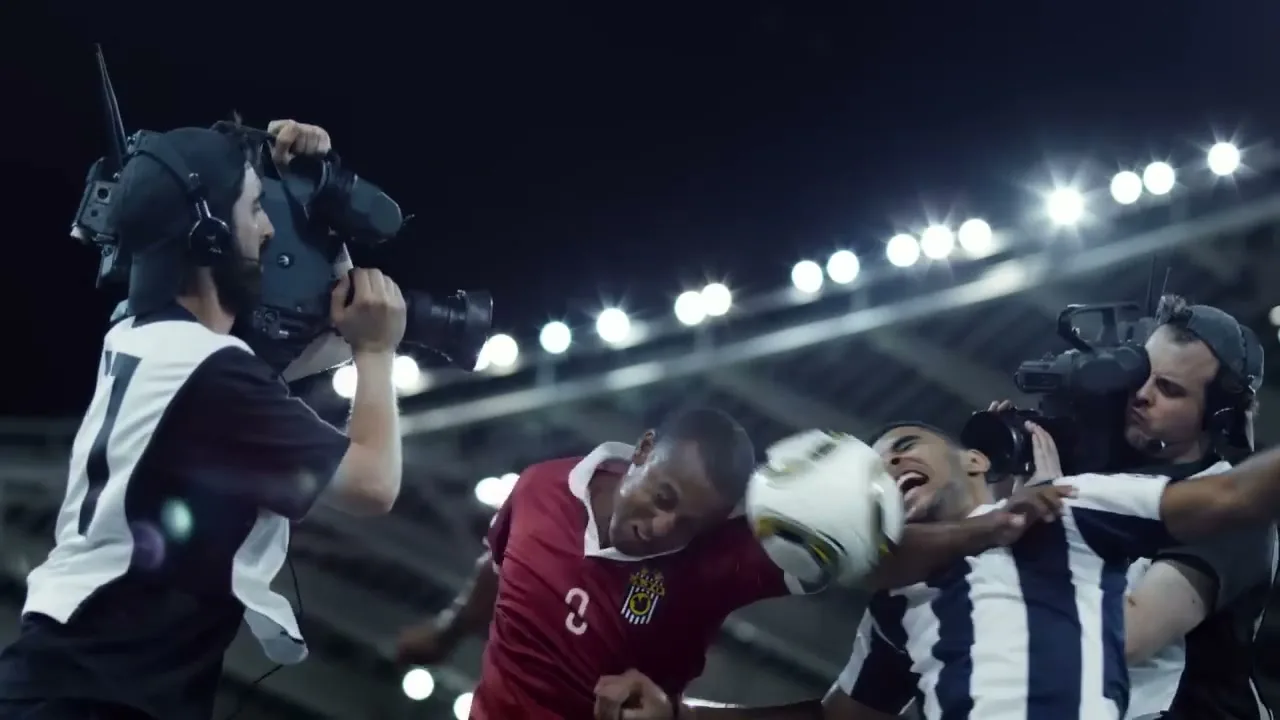 Two cameramen film a soccer match as two players headbutt a soccer ball in a stadium at night.