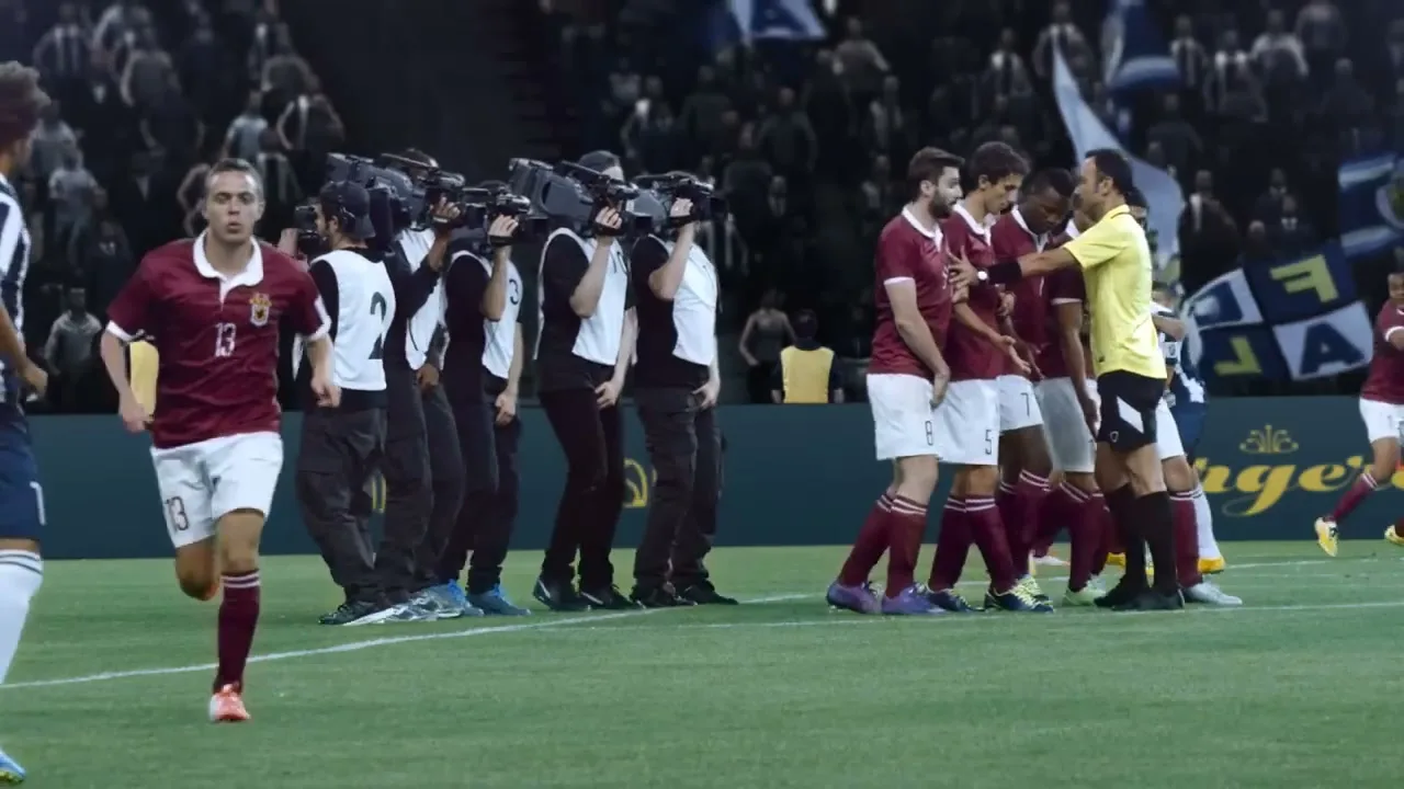 A soccer player in a maroon and white uniform runs across a green field while a line of cameramen film a confrontation between players and a referee in a stadium.