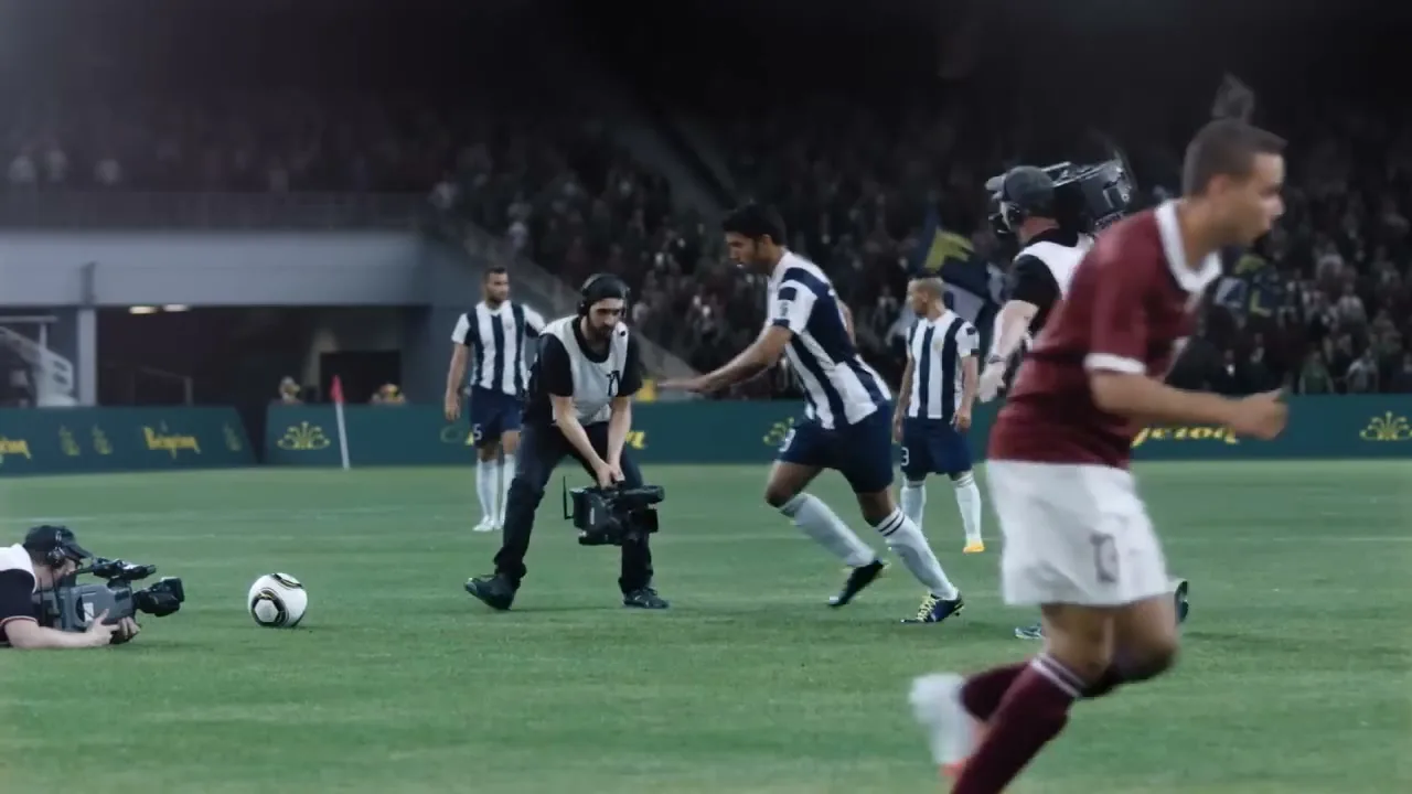 A soccer player in a maroon and white uniform runs across a green field, with other players and cameramen in the background during a game.