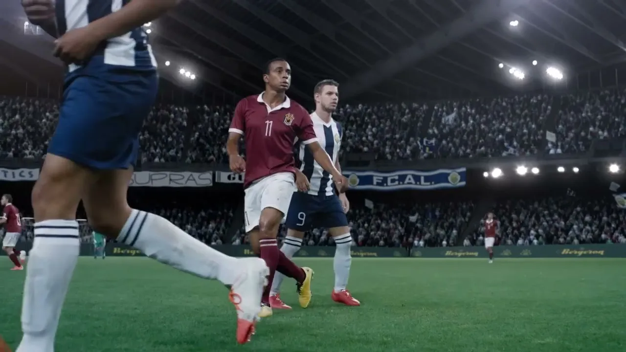 A soccer player in a maroon jersey and white shorts walks on a green field, with other players and a stadium crowd in the background.