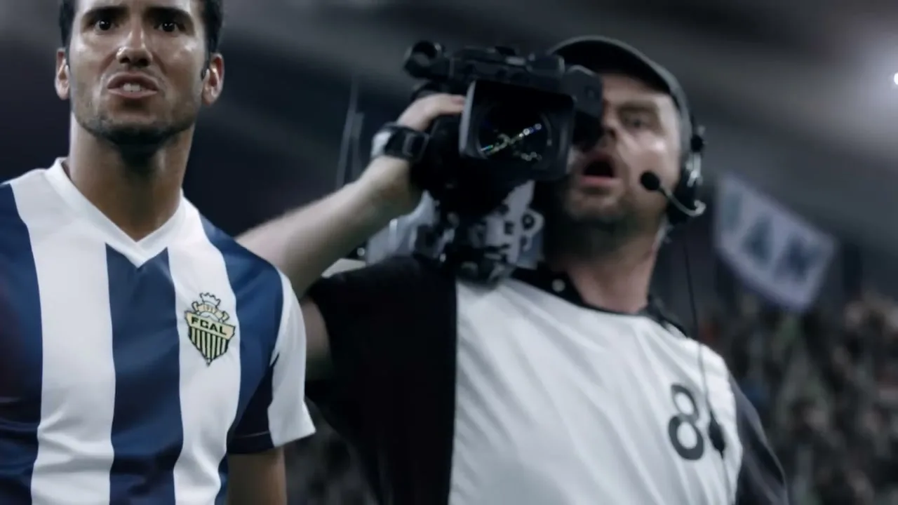 A male soccer player in a blue and white striped jersey stands next to a male cameraman with a headset, both looking intently at something off-screen in a stadium.