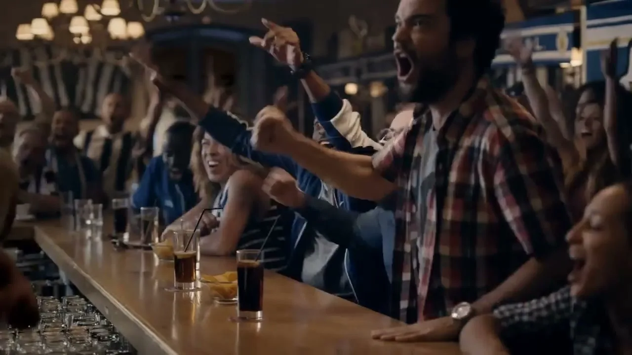 A diverse group of excited people, including men and women, cheer loudly while watching a game at a bar, with drinks and snacks on the counter.