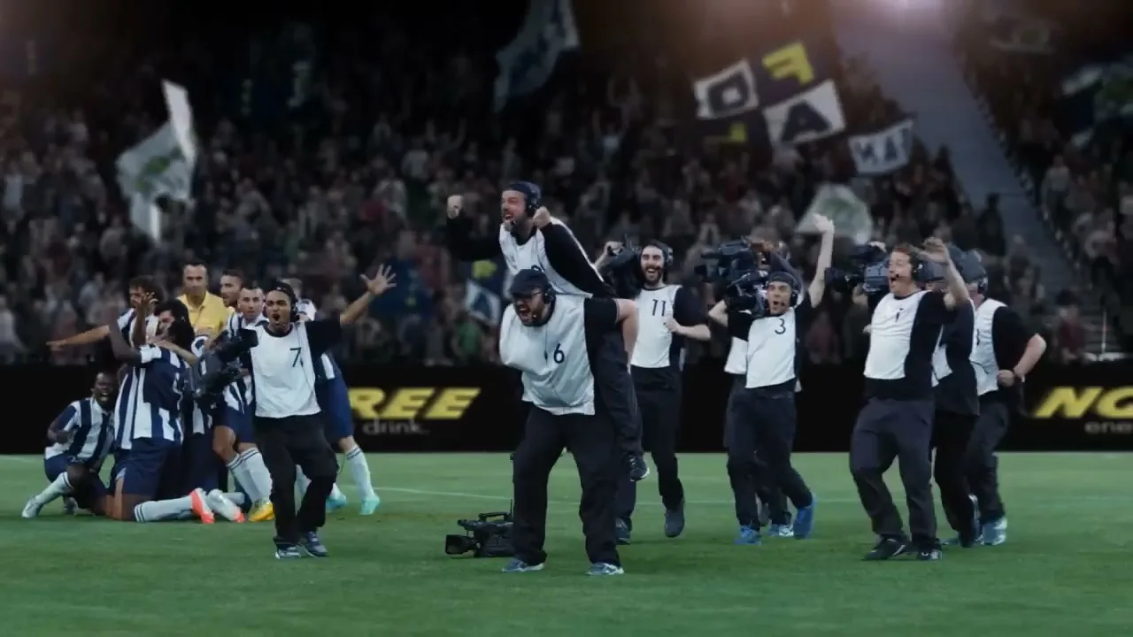 A group of cameramen in black and white uniforms celebrate with soccer players on a stadium field, with a cheering crowd in the background.