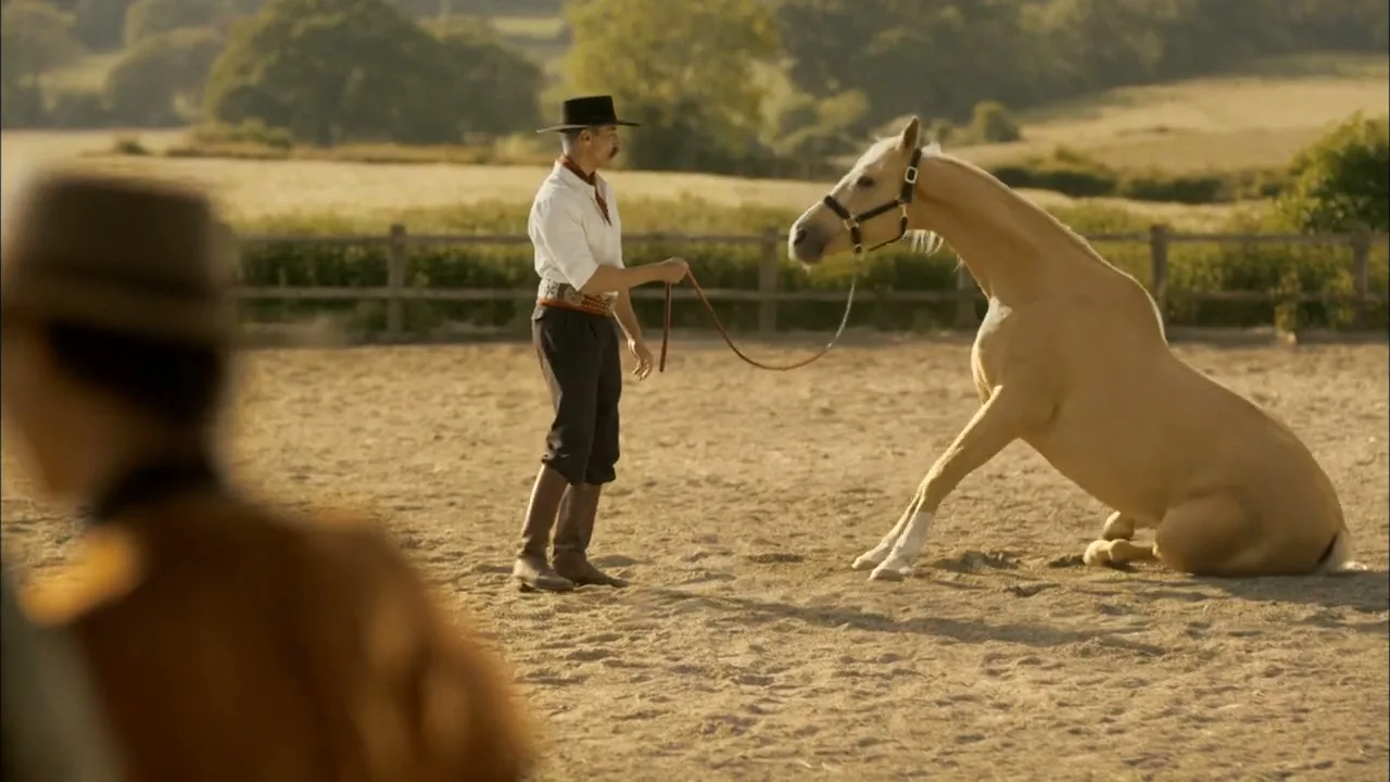 A man in a cowboy hat and traditional attire stands in a dusty arena, holding a lead rope as a palomino horse sits on the ground.