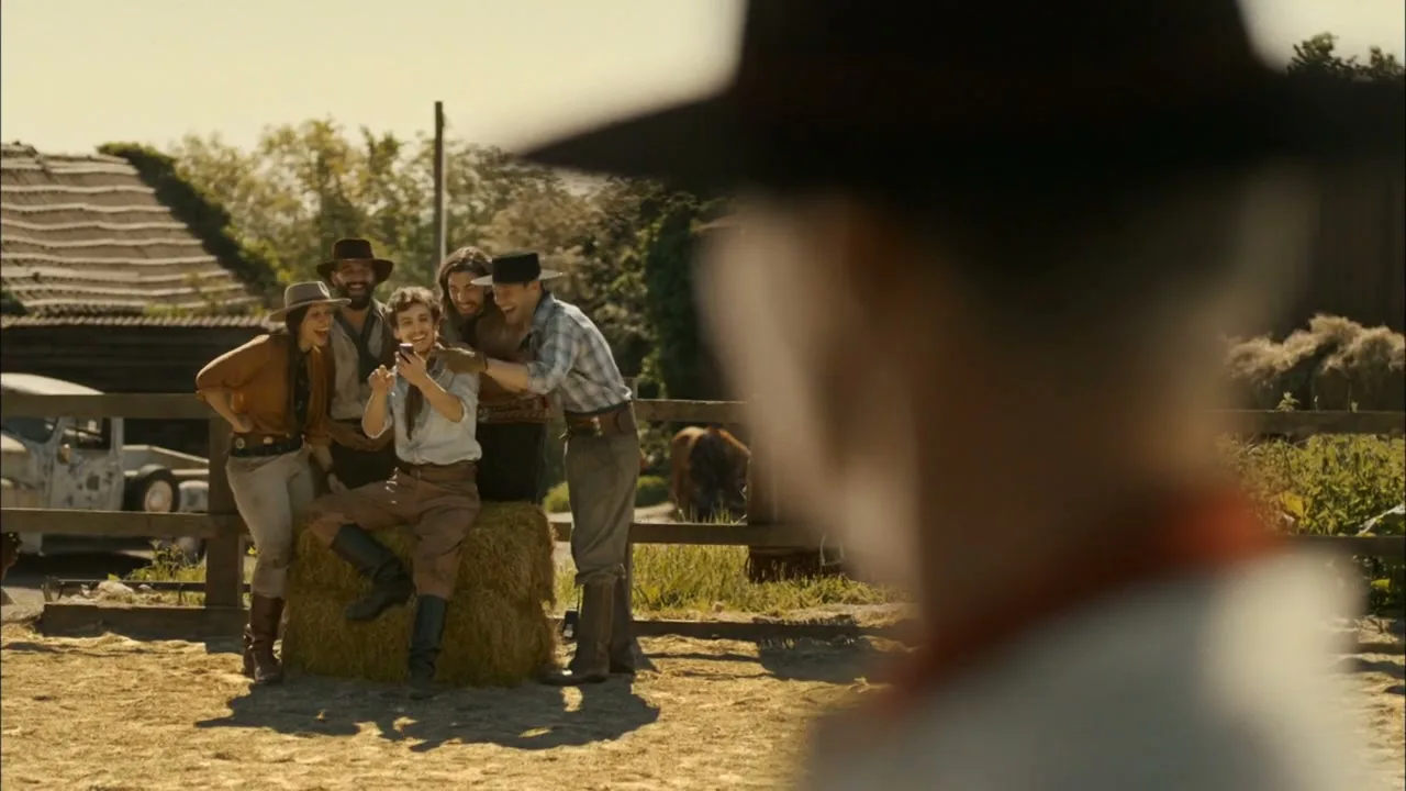 A group of five friends, dressed in cowboy attire, gather around a woman sitting on hay bales, looking at her phone and laughing, on a sunny ranch.
