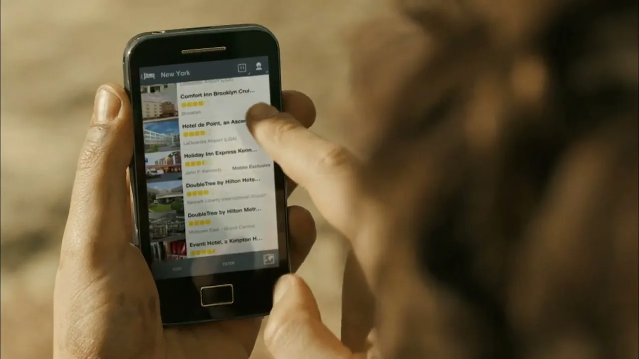 A person's hands hold a smartphone, scrolling through a list of hotels in New York on the Expedia mobile app.