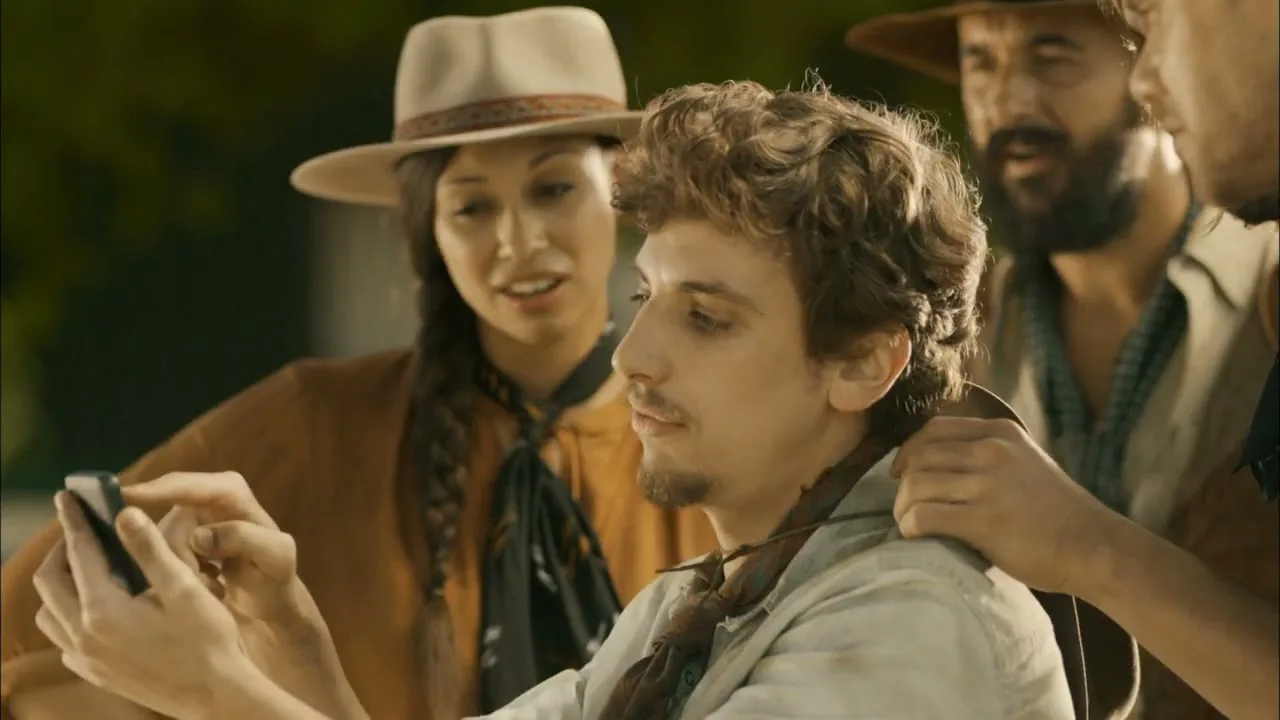 A young man in a cowboy hat and scarf looks at his phone, surrounded by three other people in cowboy attire, outdoors during the day.