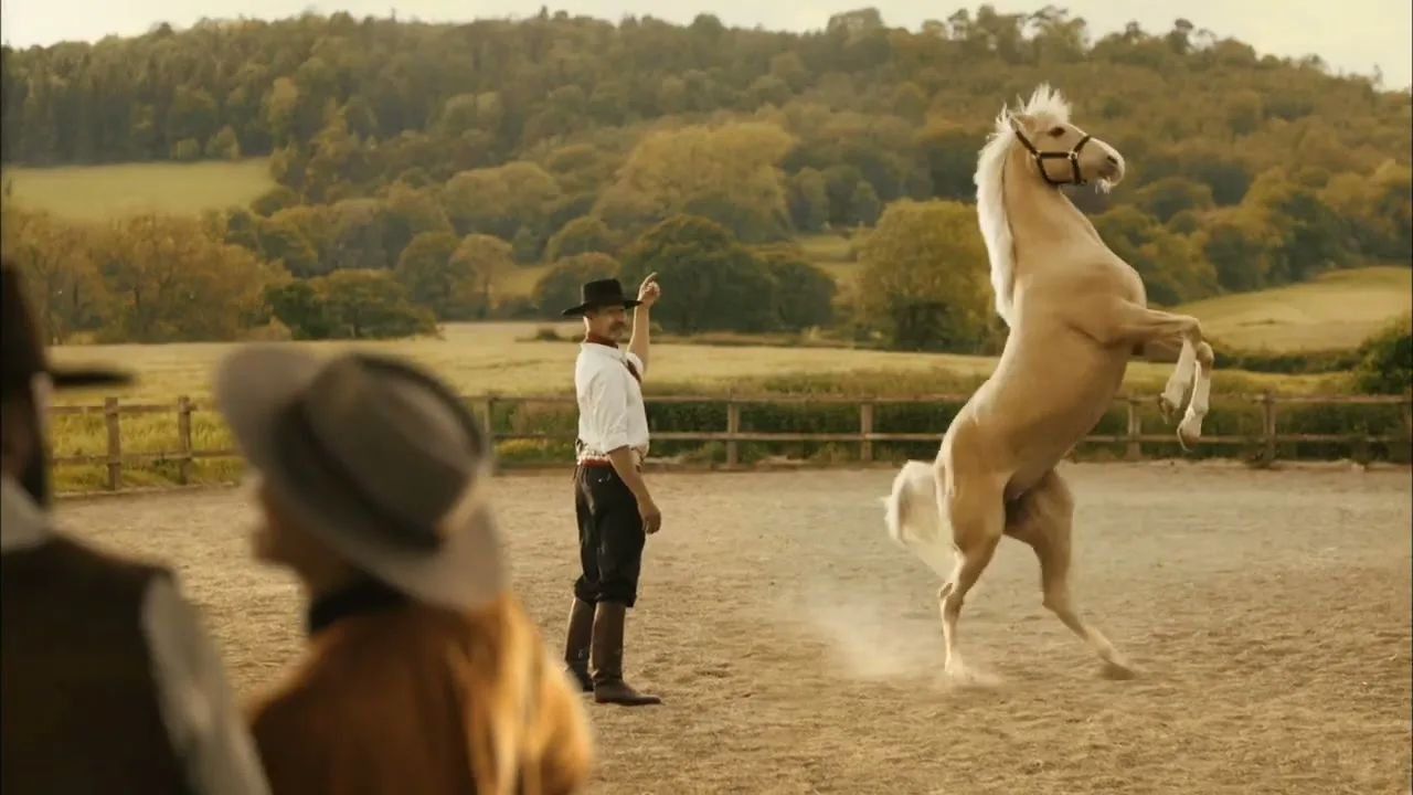 A male horse trainer in a cowboy hat and western attire stands in a dirt arena, gesturing as a palomino horse rears up on its hind legs, with two blurred spectators in the foreground.