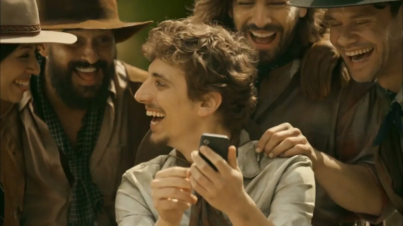 A young man with curly hair smiles and laughs while looking at his phone, surrounded by a group of laughing people in cowboy attire.
