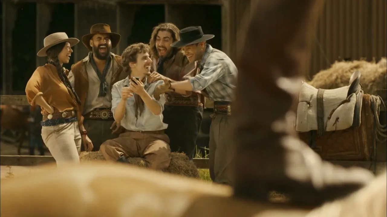 A group of five people, dressed in cowboy attire, laugh and interact with a man holding a phone, in a ranch setting.