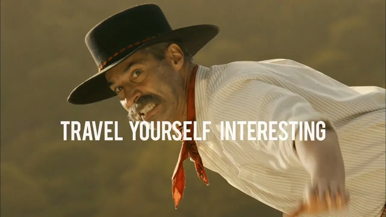 A man with a mustache and a cowboy hat leans forward, looking intensely at the camera with a slight smile, in an outdoor setting.