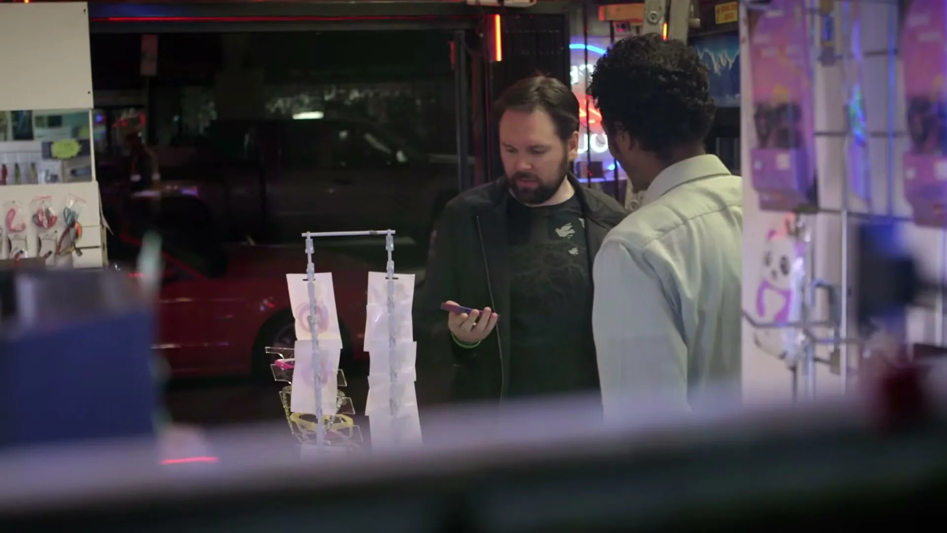 Two men, one with a beard and dark clothing, the other with curly hair and a light shirt, talk in a brightly lit electronics store at night.