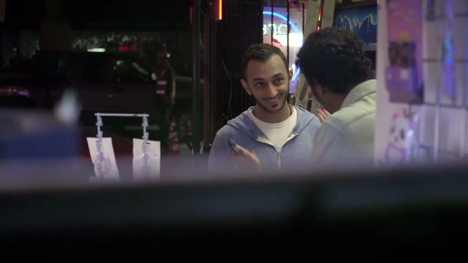 Two men are talking to each other in a store at night, with neon signs visible in the background.