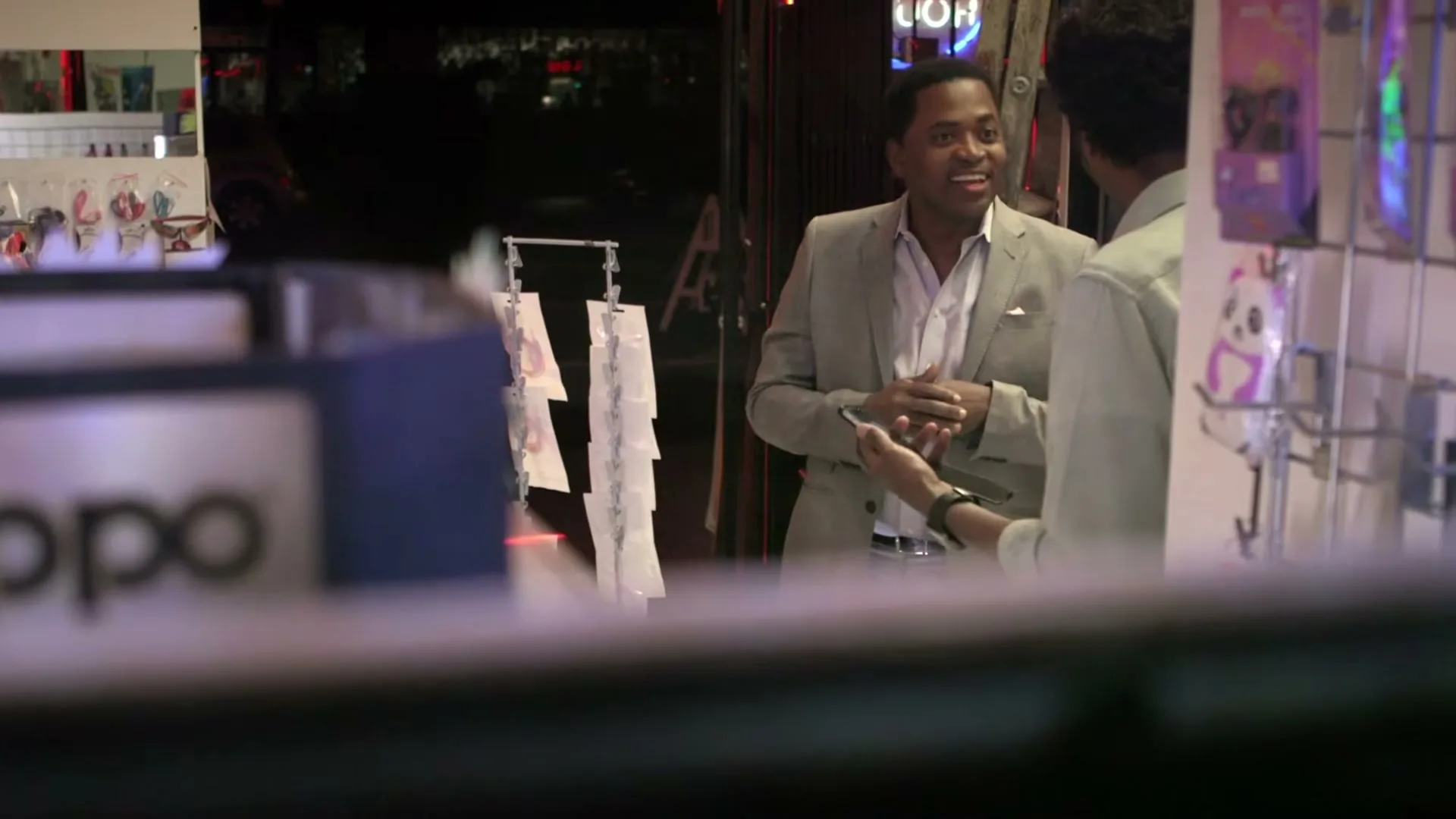 Two men, one in a suit and one in a light shirt, are talking and smiling in a brightly lit store at night.