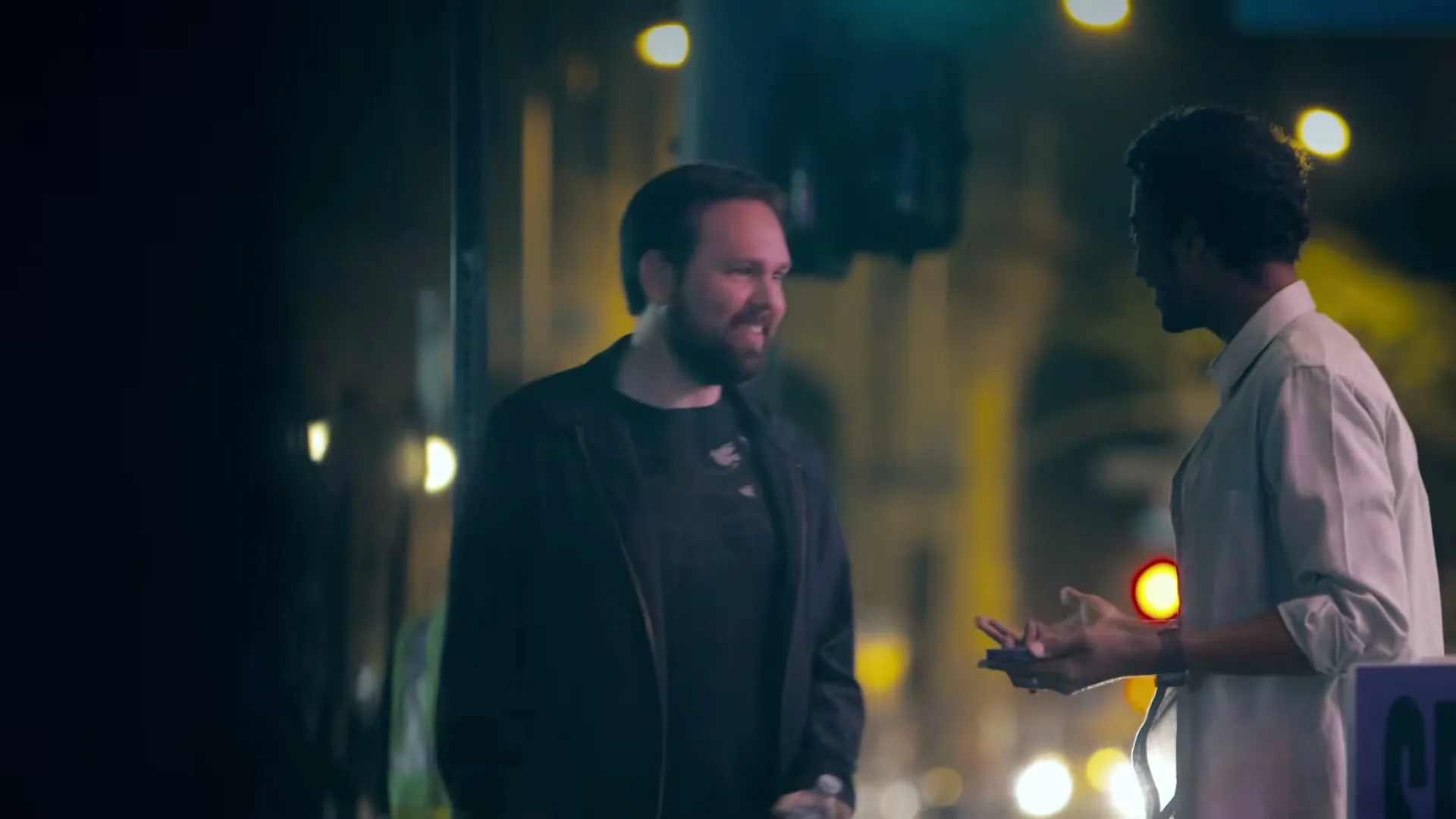 Two men, one with a beard in a black jacket and another in a light shirt, stand talking on a city street at night, with blurred lights in the background.