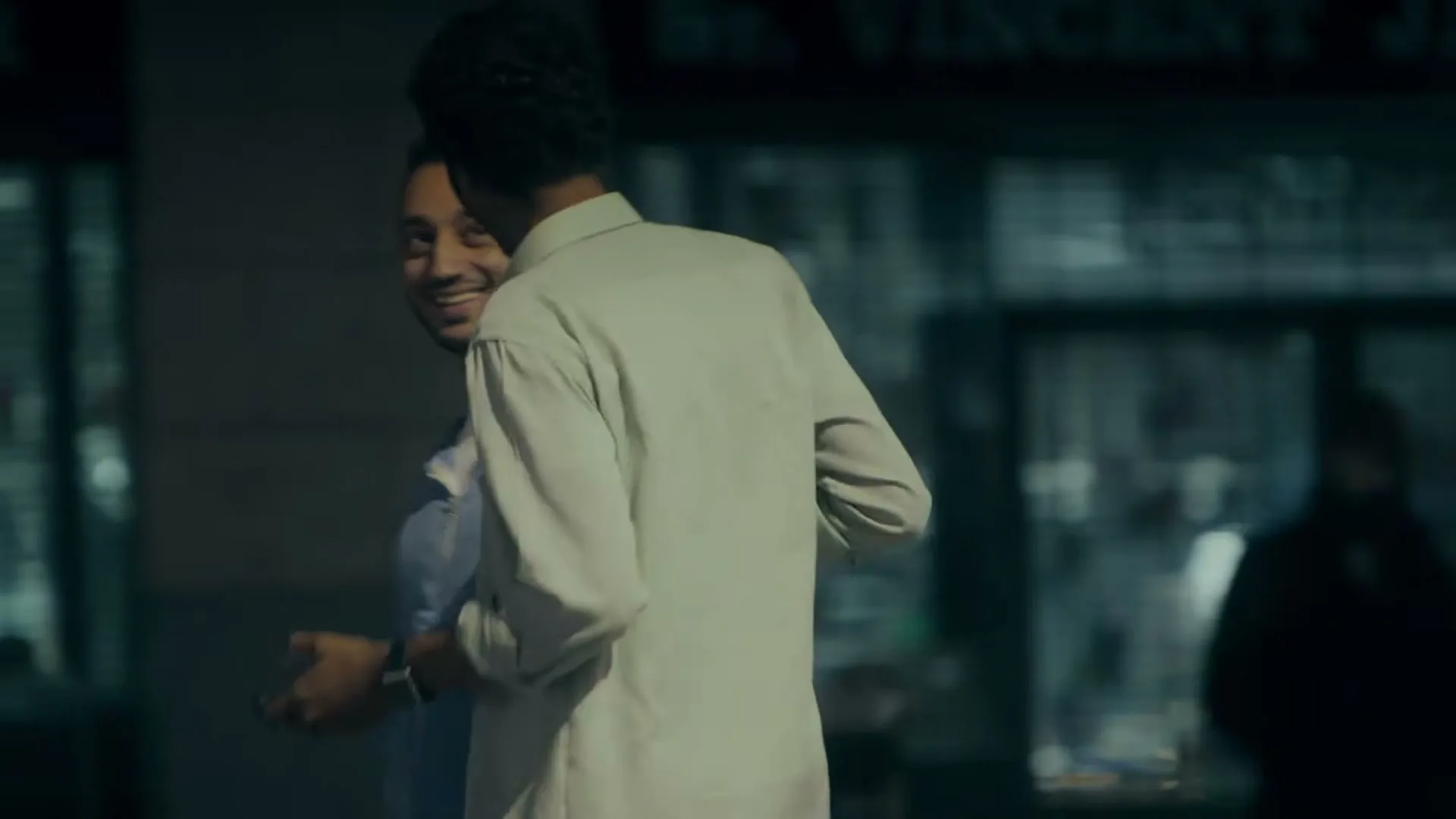 Two men, one in a light blue hoodie and the other in a light-colored button-up shirt, are smiling and interacting on a city street at night.