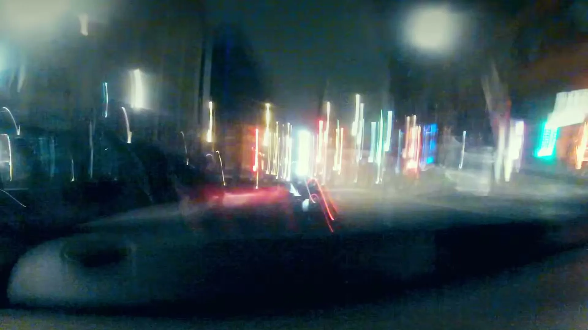 A POV shot from inside a car shows a red car driving past on a busy city street at night, with bright lights and buildings in the background.