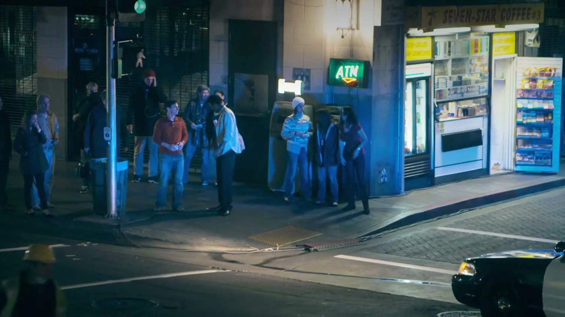 A diverse group of people stand on a city sidewalk at night, near a convenience store and an ATM, as a police car drives by.