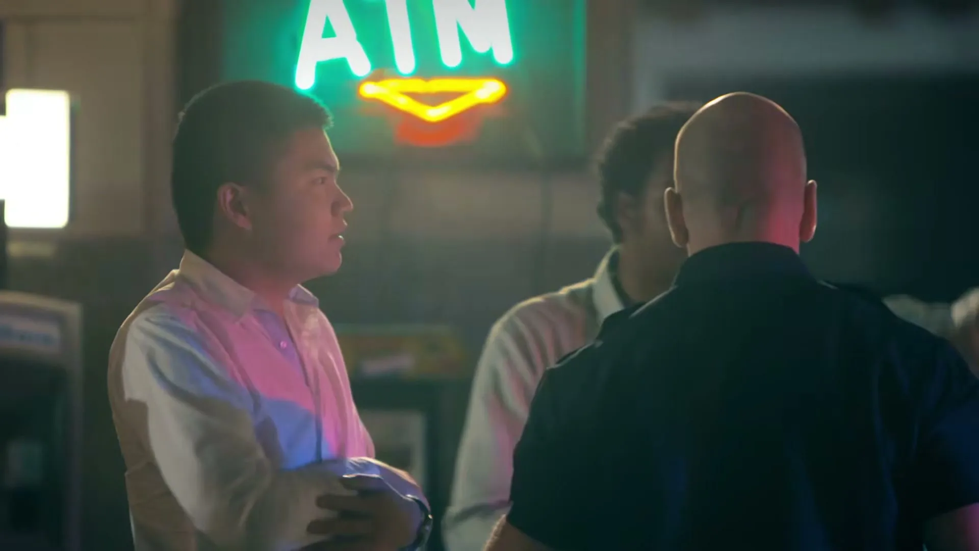 Three men, one in a white shirt, one in a dark shirt, and one with curly hair, stand near a neon ATM sign at night.