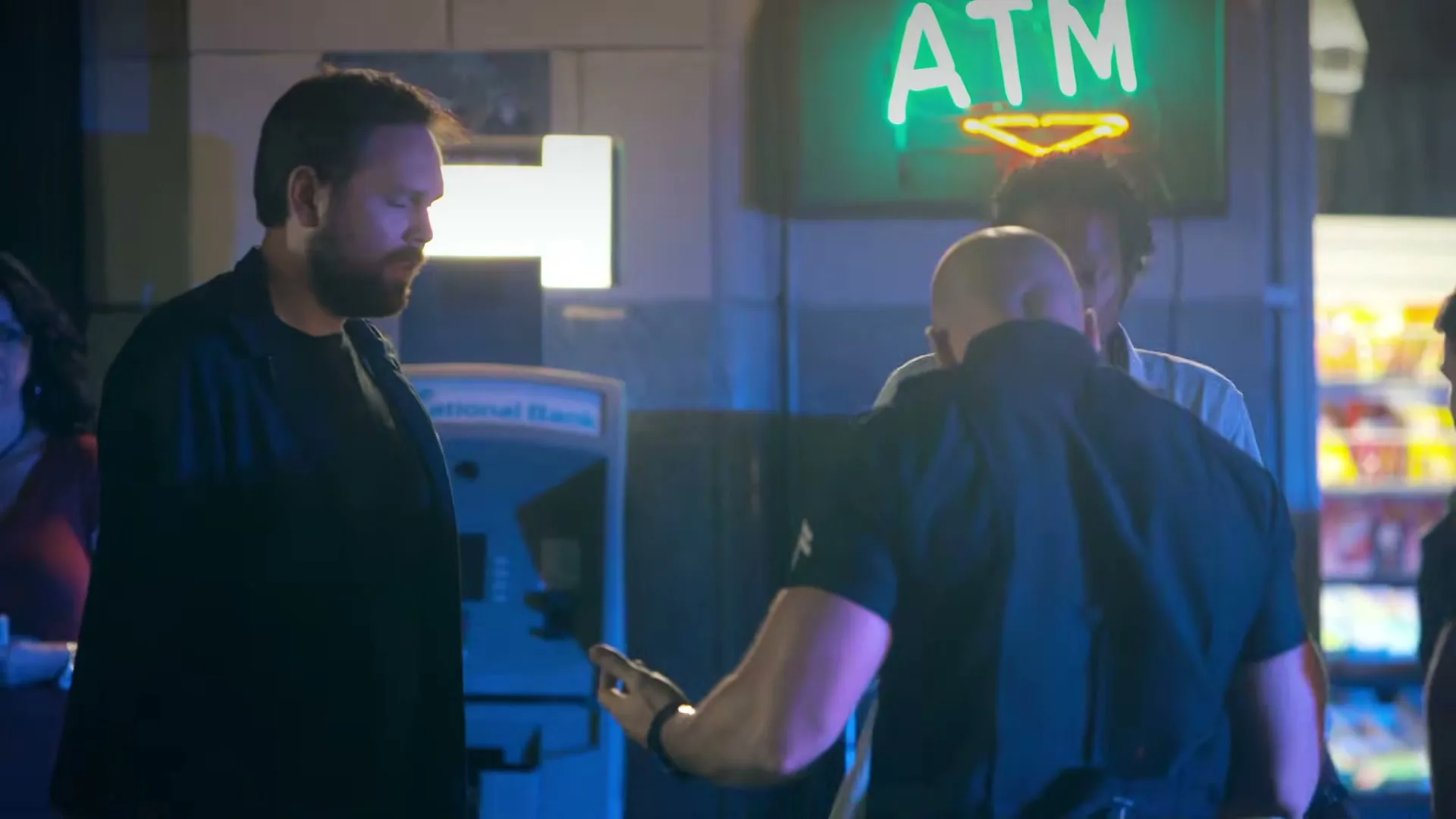 A man with a beard looks on as a bald man in a black shirt talks to another man in front of an ATM at night.
