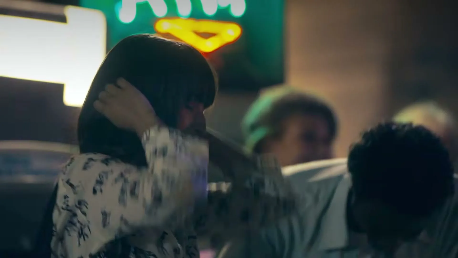A young woman with dark hair in a patterned shirt covers her ears, then smiles, next to a man clapping, under a neon ATM sign at night.