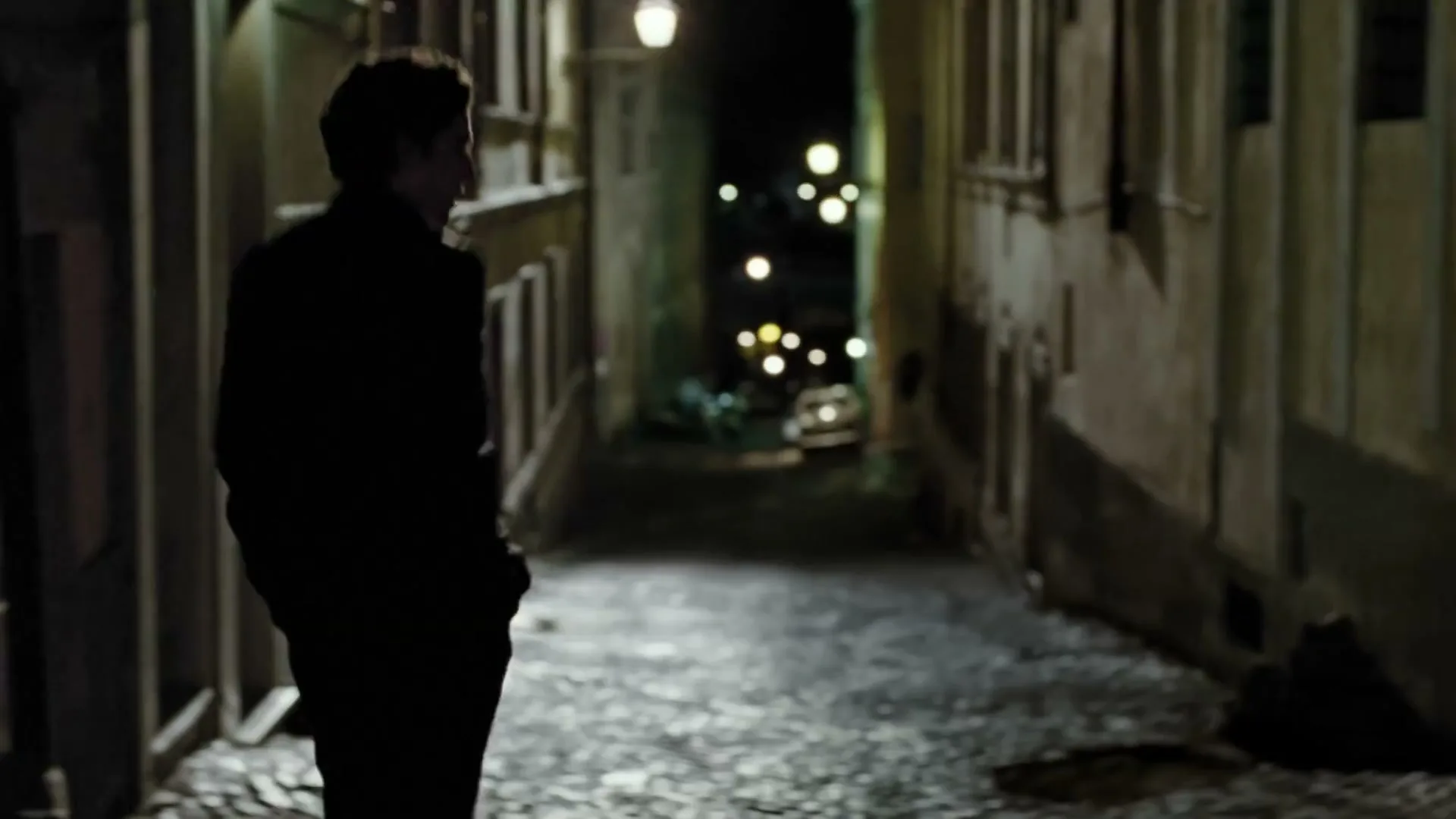 A man with dark curly hair, wearing a suit, looks up with a pensive expression in a dark, narrow alleyway at night.