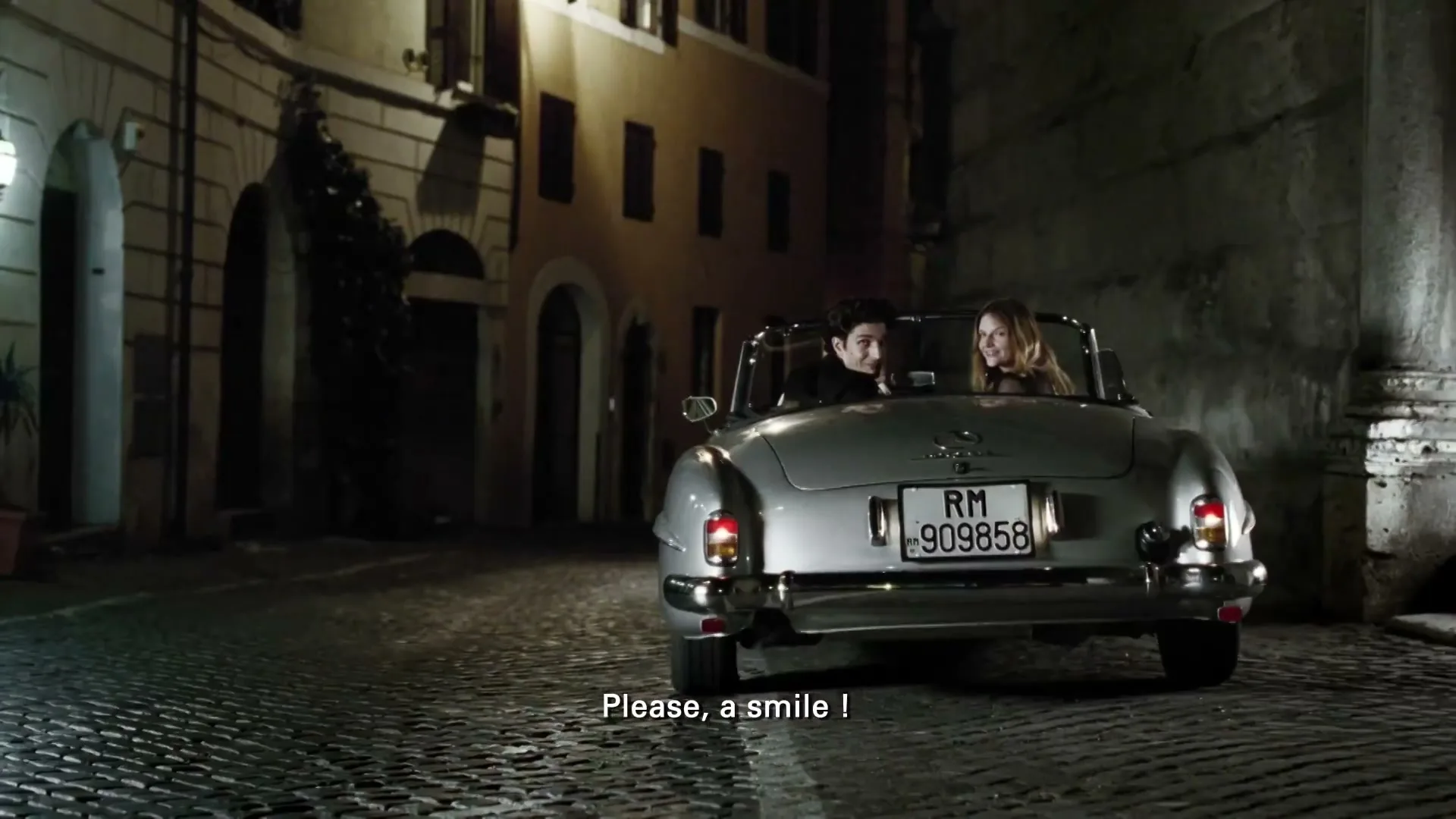 A man and woman in a vintage silver convertible car are parked on a cobblestone street at night in an old European city.