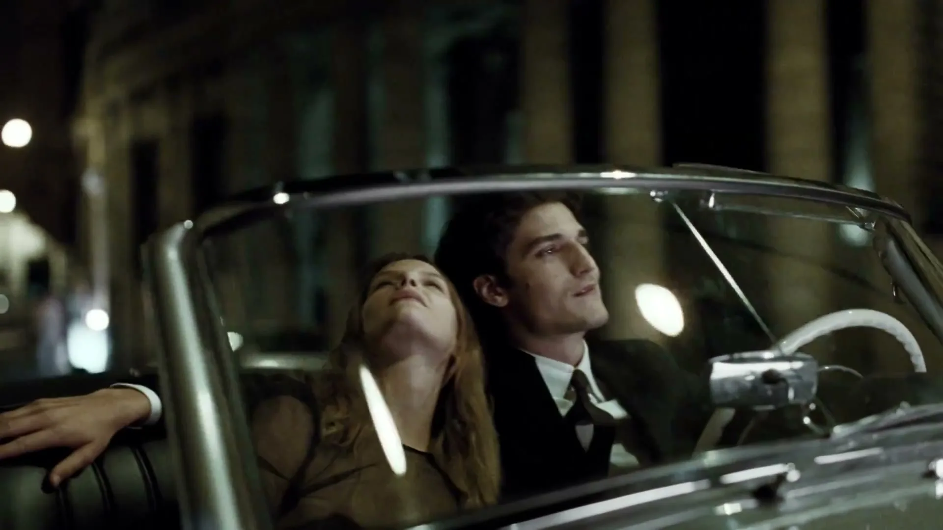 A man in a suit drives a convertible at night, while a woman with long hair leans back in the passenger seat, looking up at the sky.