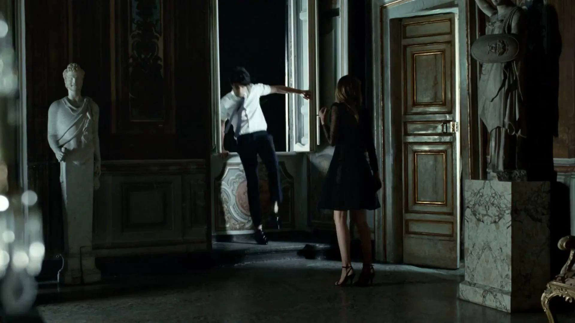A man in a white shirt and dark pants climbs through a window while a woman in a black dress watches him in a dimly lit, ornate room.