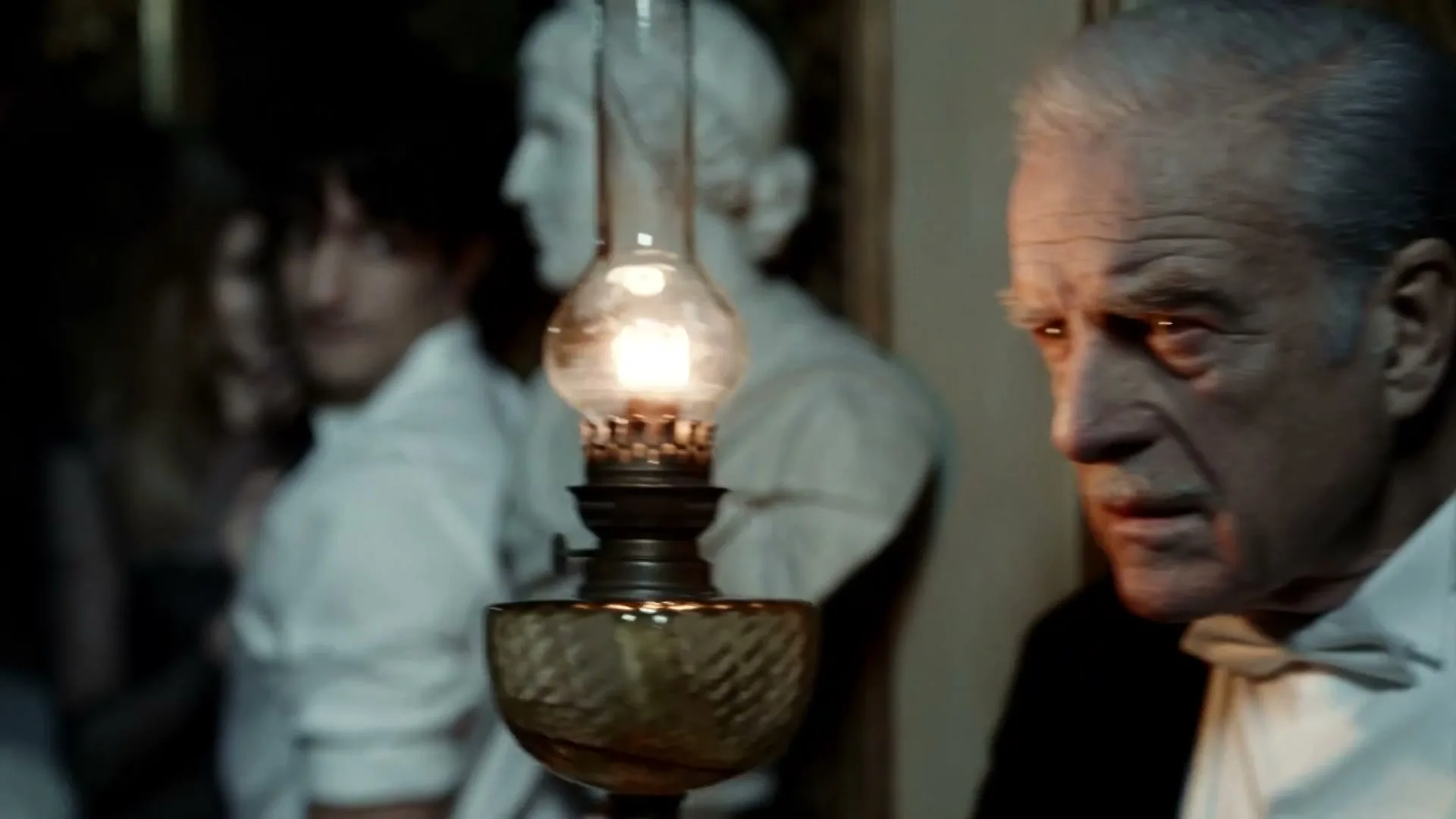 An older man in a tuxedo looks intently at something off-screen, illuminated by a flickering oil lamp in a dimly lit, ornate interior.