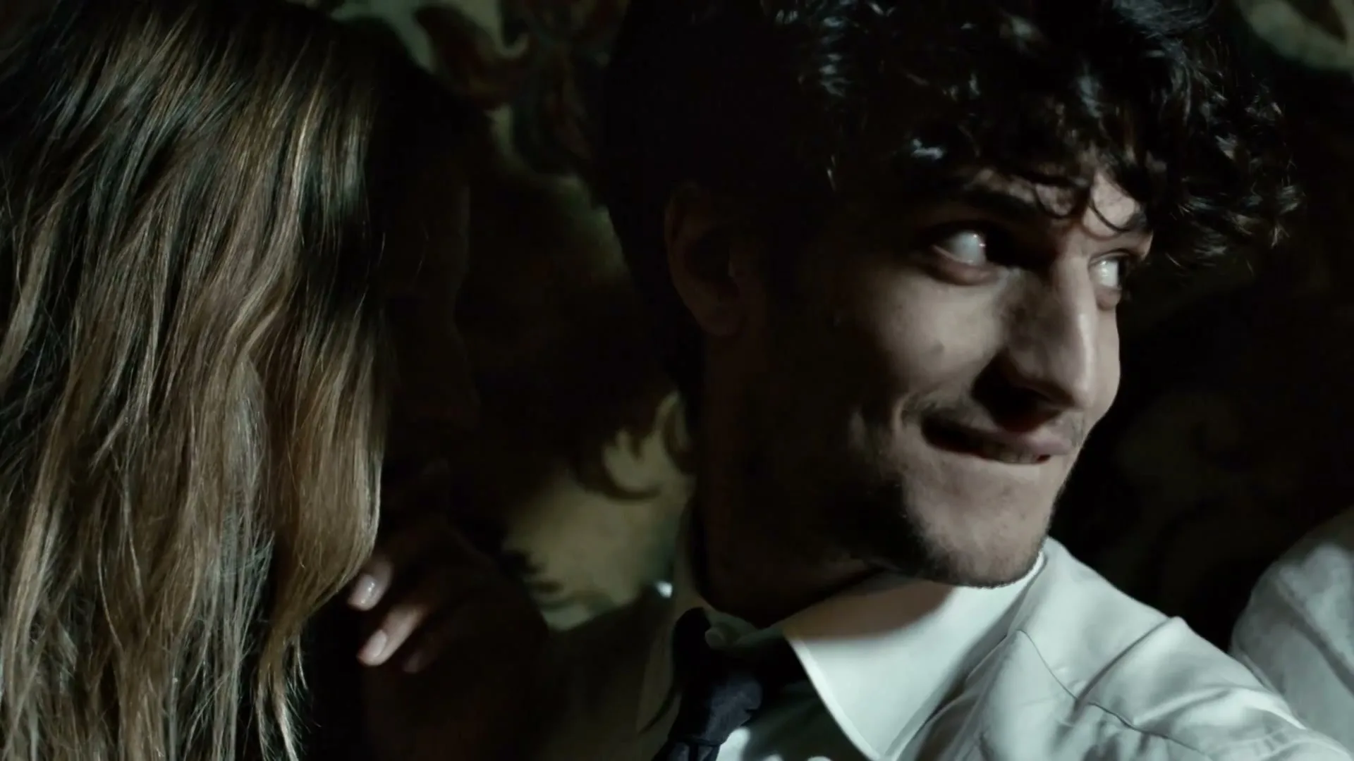 A man with curly hair and a white shirt looks sideways with a smirk, while a woman's hair and hand are visible next to him in a dimly lit interior.