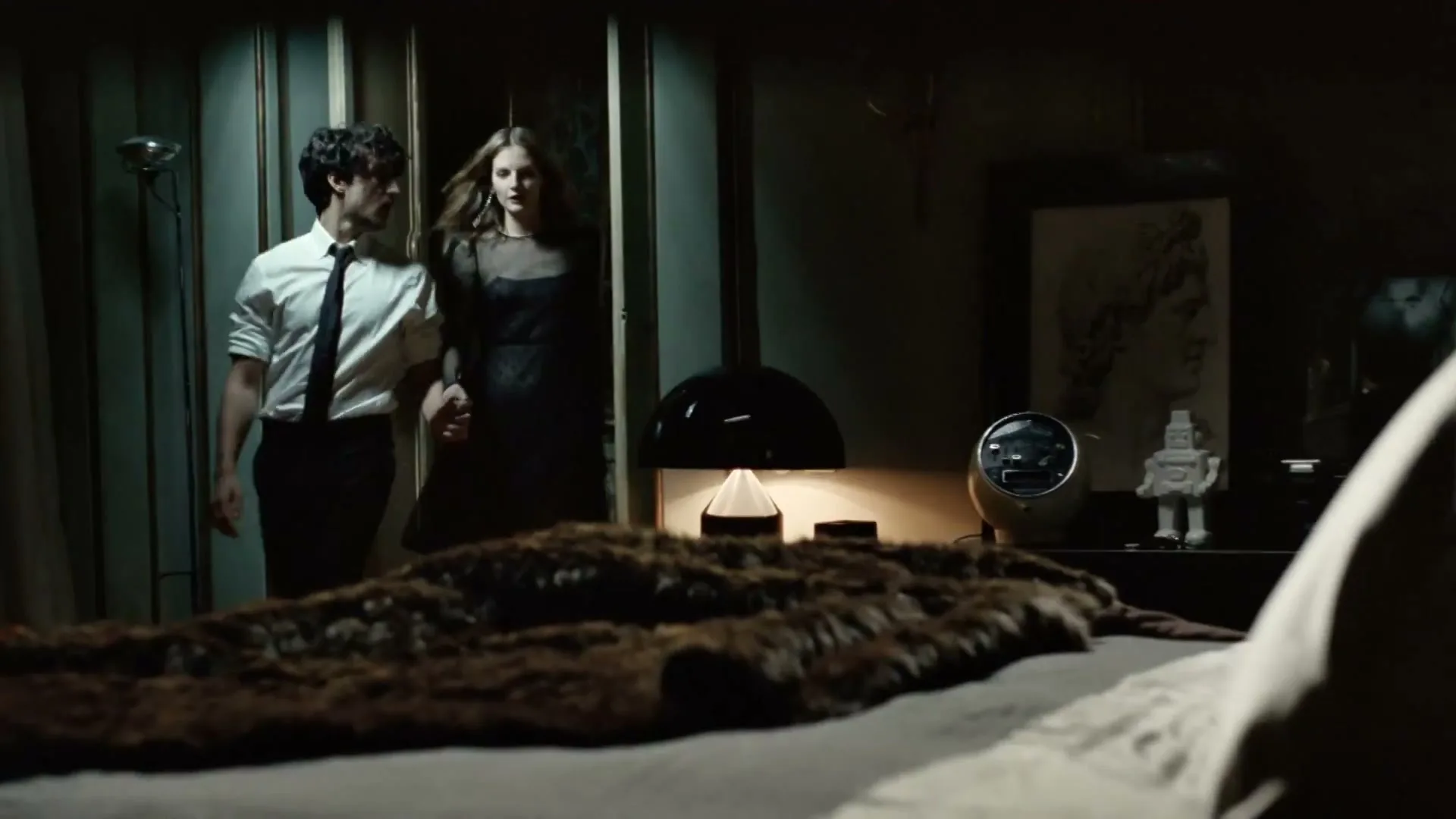 A man and woman, dressed in formal attire, walk into a dimly lit bedroom, holding hands.