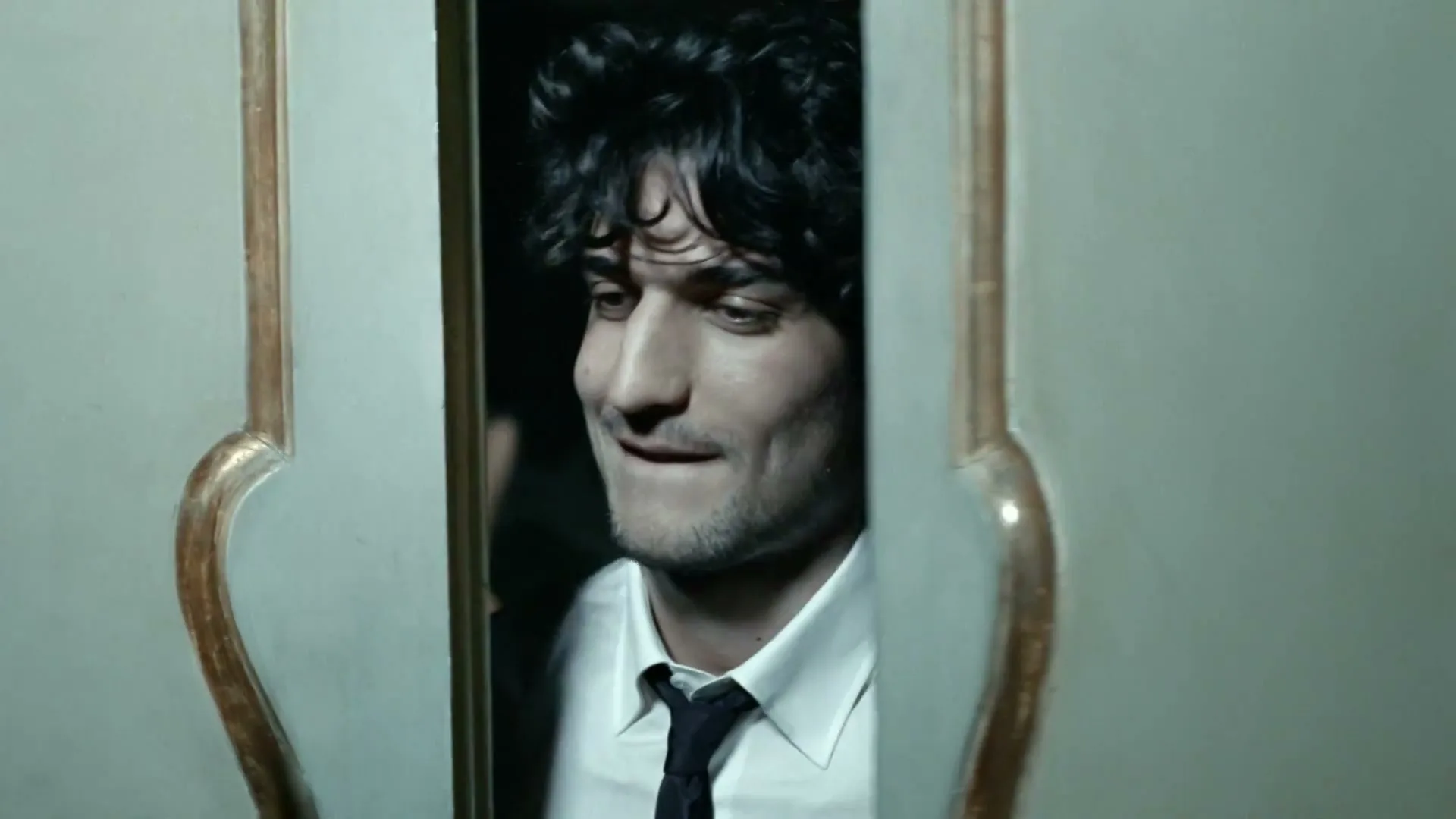 A young man with dark curly hair, wearing a white shirt and black tie, looks around nervously from behind a partially open door.