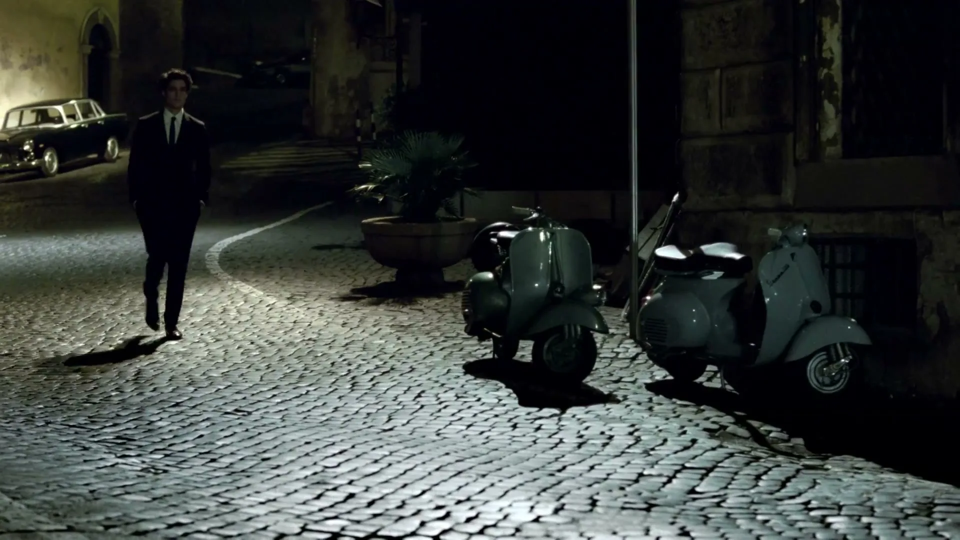 A man in a suit walks on a cobblestone street at night, with two scooters parked nearby and a classic car in the background.