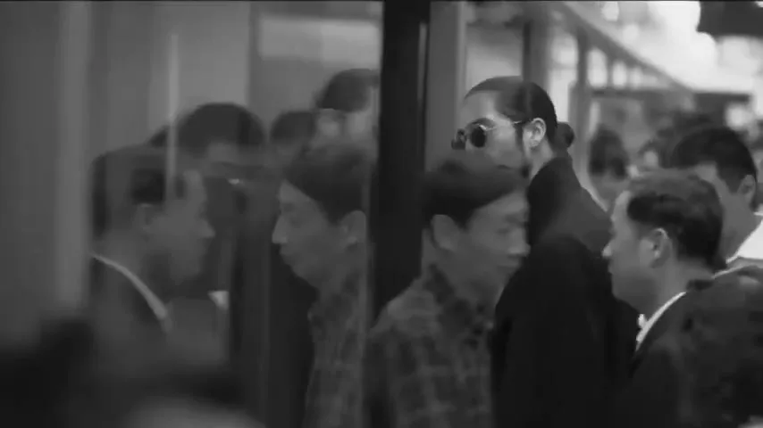 A man with a beard and sunglasses walks through a crowded indoor space, looking to his left, with reflections visible in a glass partition.