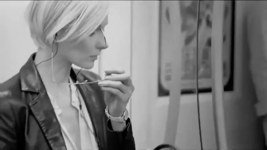 A stylish woman with short blonde hair takes off her glasses while sitting on a train, looking down at a newspaper.