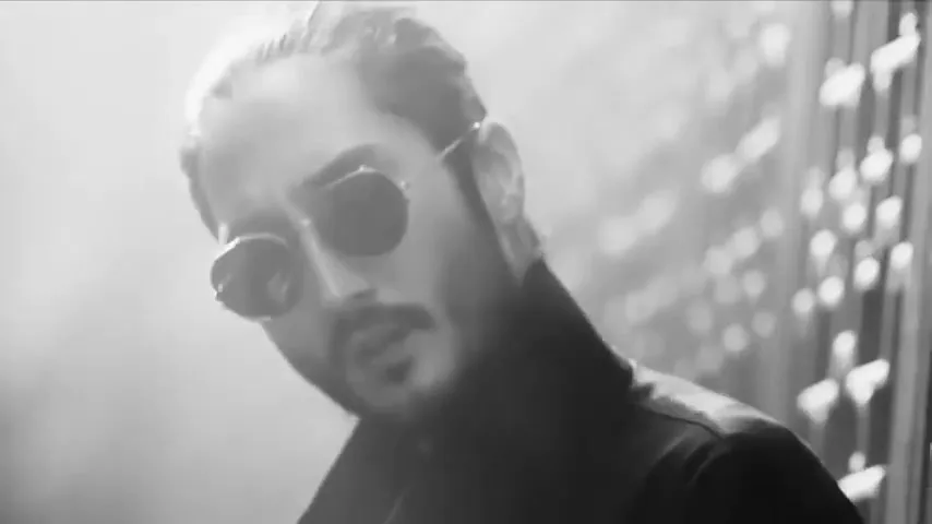 A man with a beard and sunglasses looks down, then up, then down again, in a black and white shot with bright light.