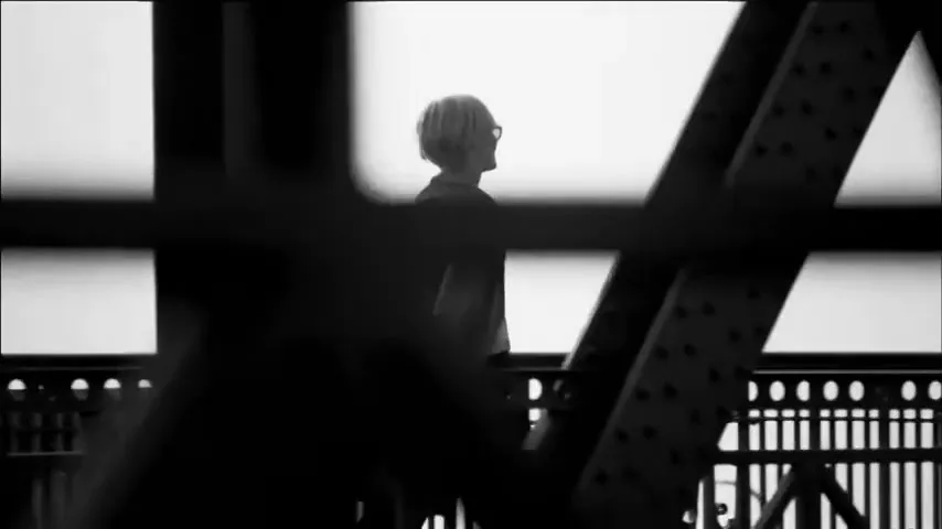 A person with short hair and glasses walks along a bridge, seen through the dark, silhouetted structure of the bridge itself.