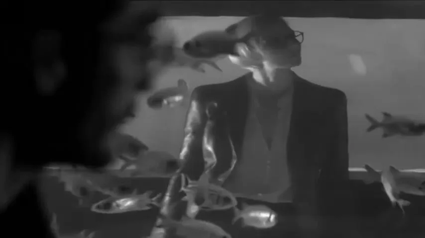 A man in glasses and a suit stands behind an aquarium, looking up and to the right, as fish swim around him.