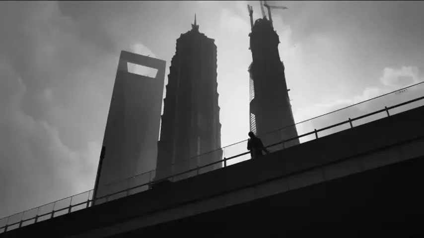 A silhouetted man walks along a bridge railing with three towering skyscrapers in the background, under a cloudy sky.