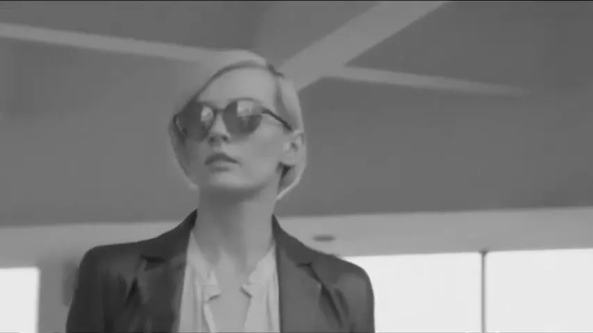 A woman with short blonde hair and sunglasses looks off-camera, wearing a blazer and blouse in a modern interior space.