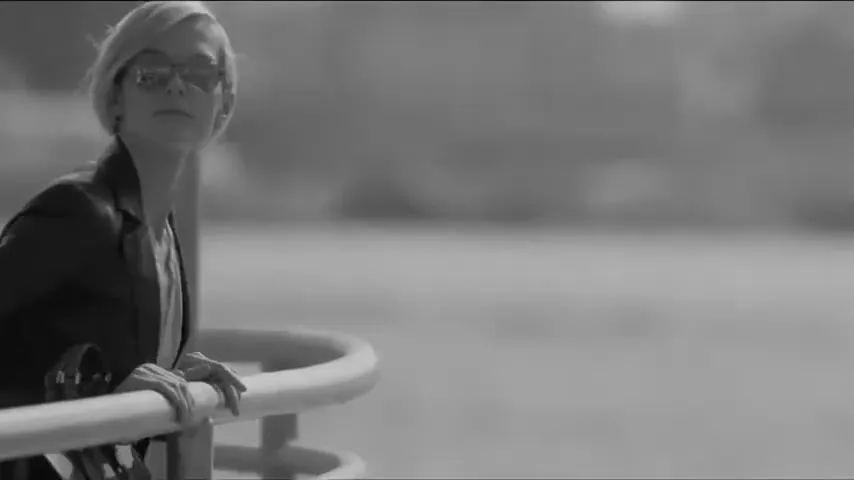 A stylish woman with short blonde hair and sunglasses leans on a railing, looking out over a body of water in black and white.