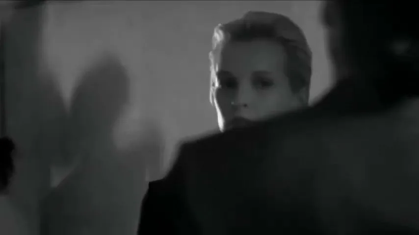 A woman with blonde hair in a dark suit looks intently at someone off-screen in a dimly lit room, with shadows on the wall.