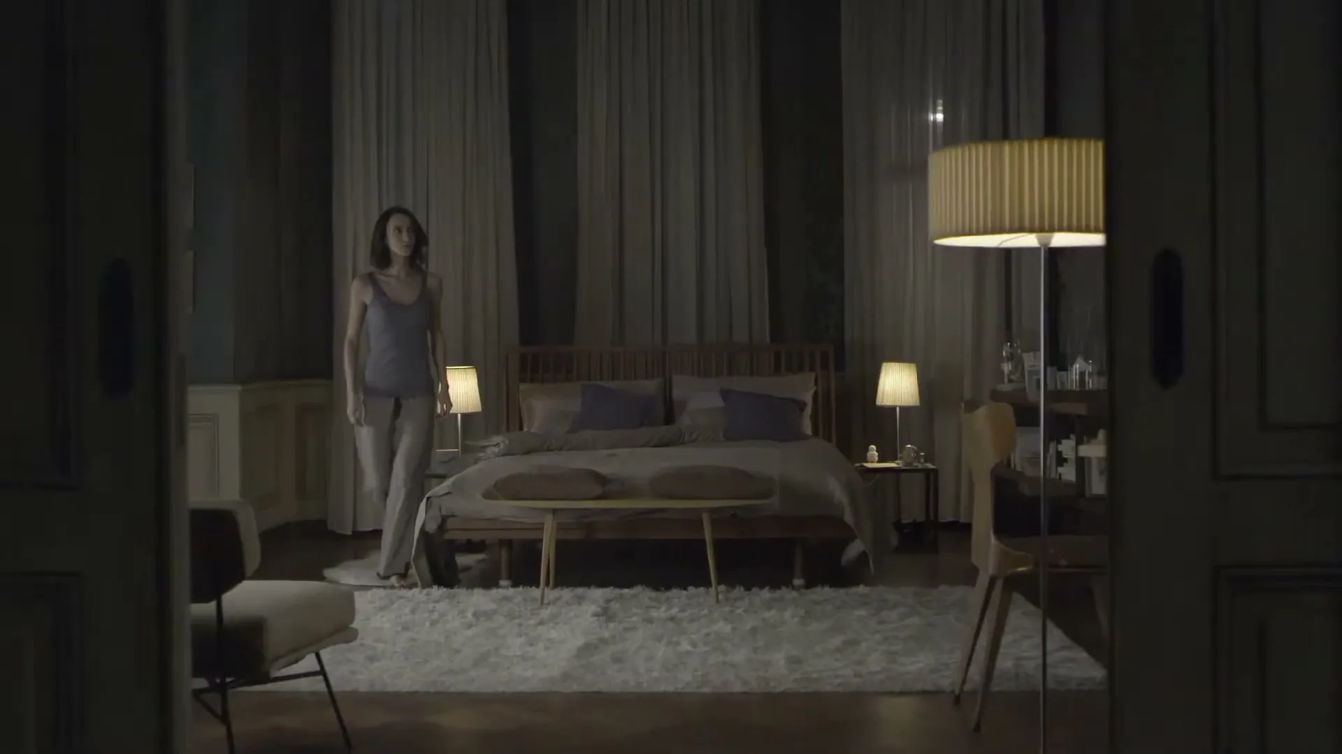 A woman in pajamas walks into a dimly lit bedroom, turning on lights as she enters, seen through an open doorway.