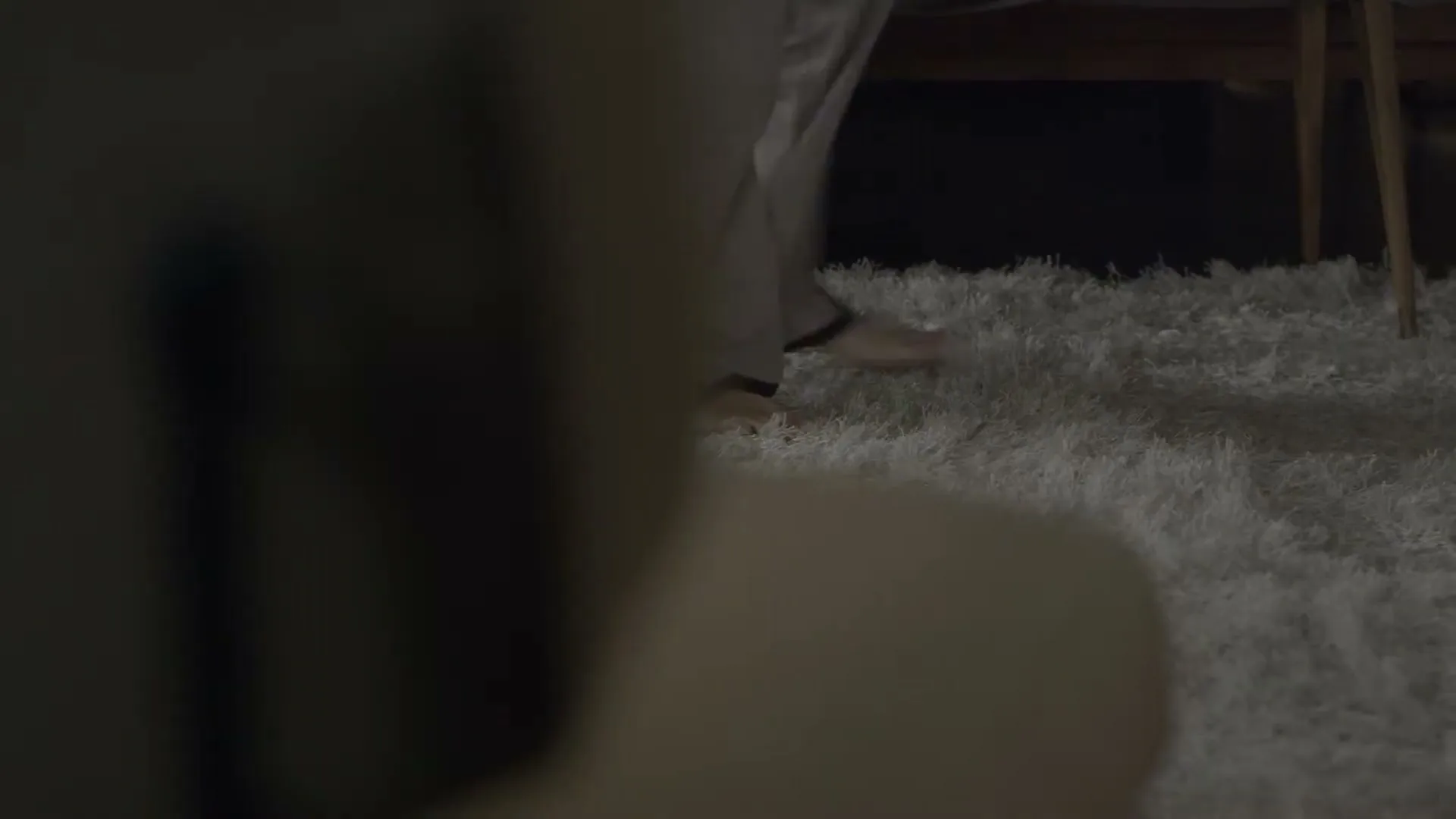 A person's bare feet and lower legs in light-colored pajamas walk across a white shag rug in a dimly lit room.