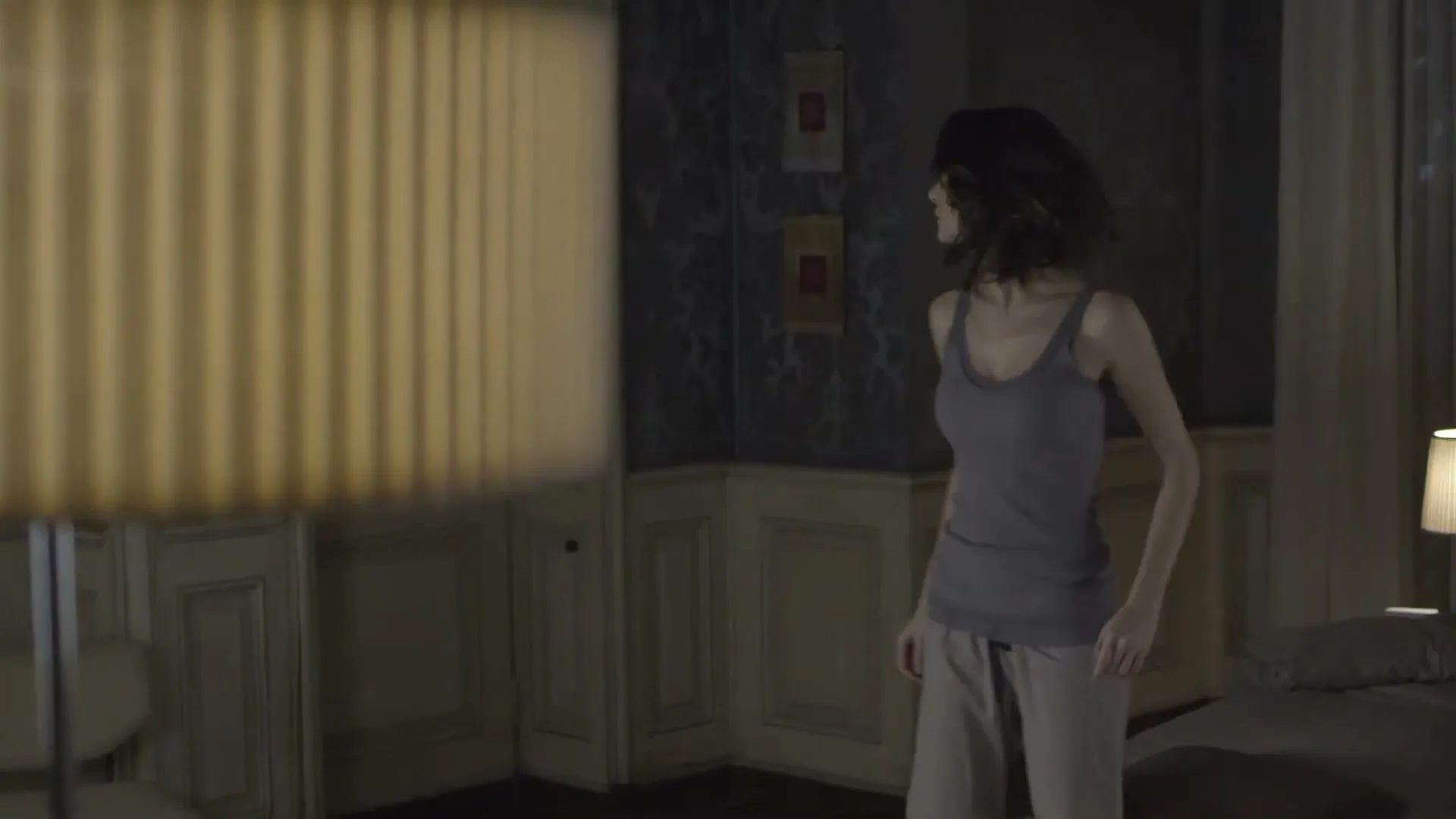 A woman in a tank top and pajama pants walks slowly through a dimly lit bedroom, looking around cautiously.