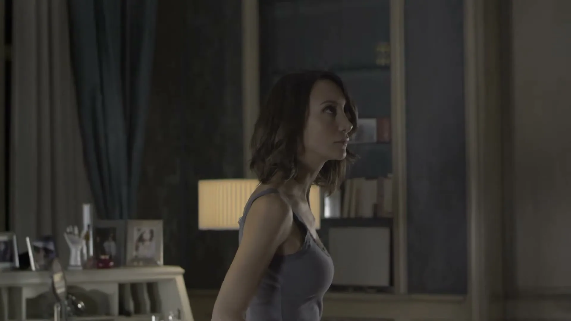 A woman with short brown hair, wearing a grey tank top, stands in a dimly lit room, looking up and to the right.