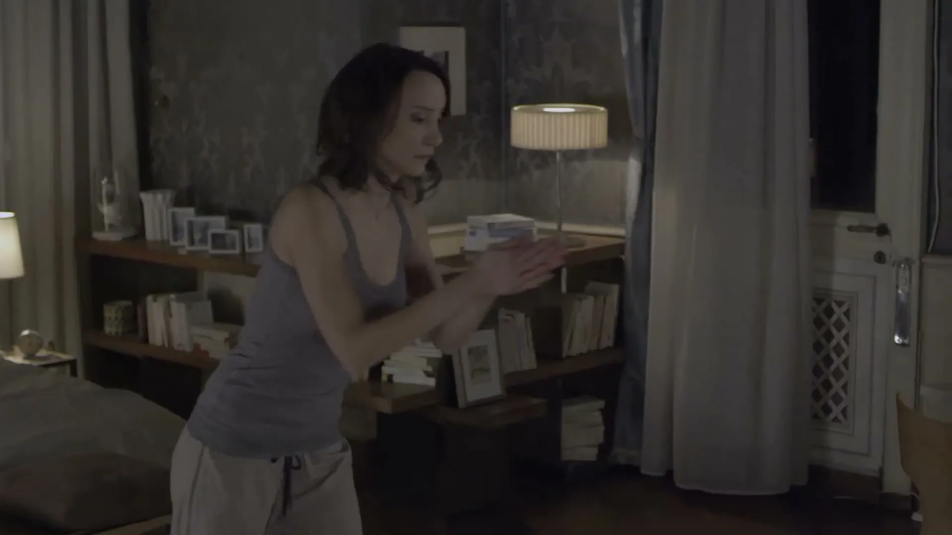 A woman in her 30s, wearing a tank top and pajama pants, claps her hands in a dimly lit bedroom.