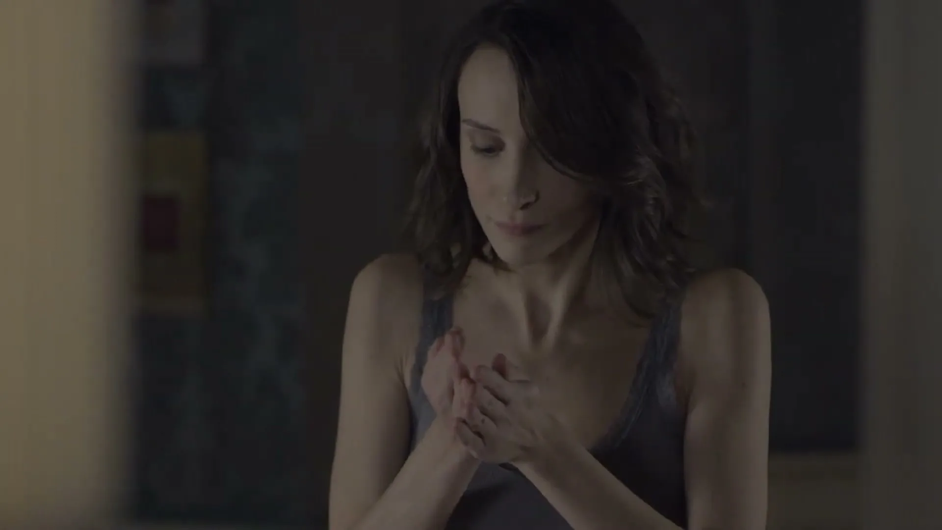 A woman with dark hair in a grey tank top rubs her hands together, then looks up with a concerned expression in a dimly lit room.