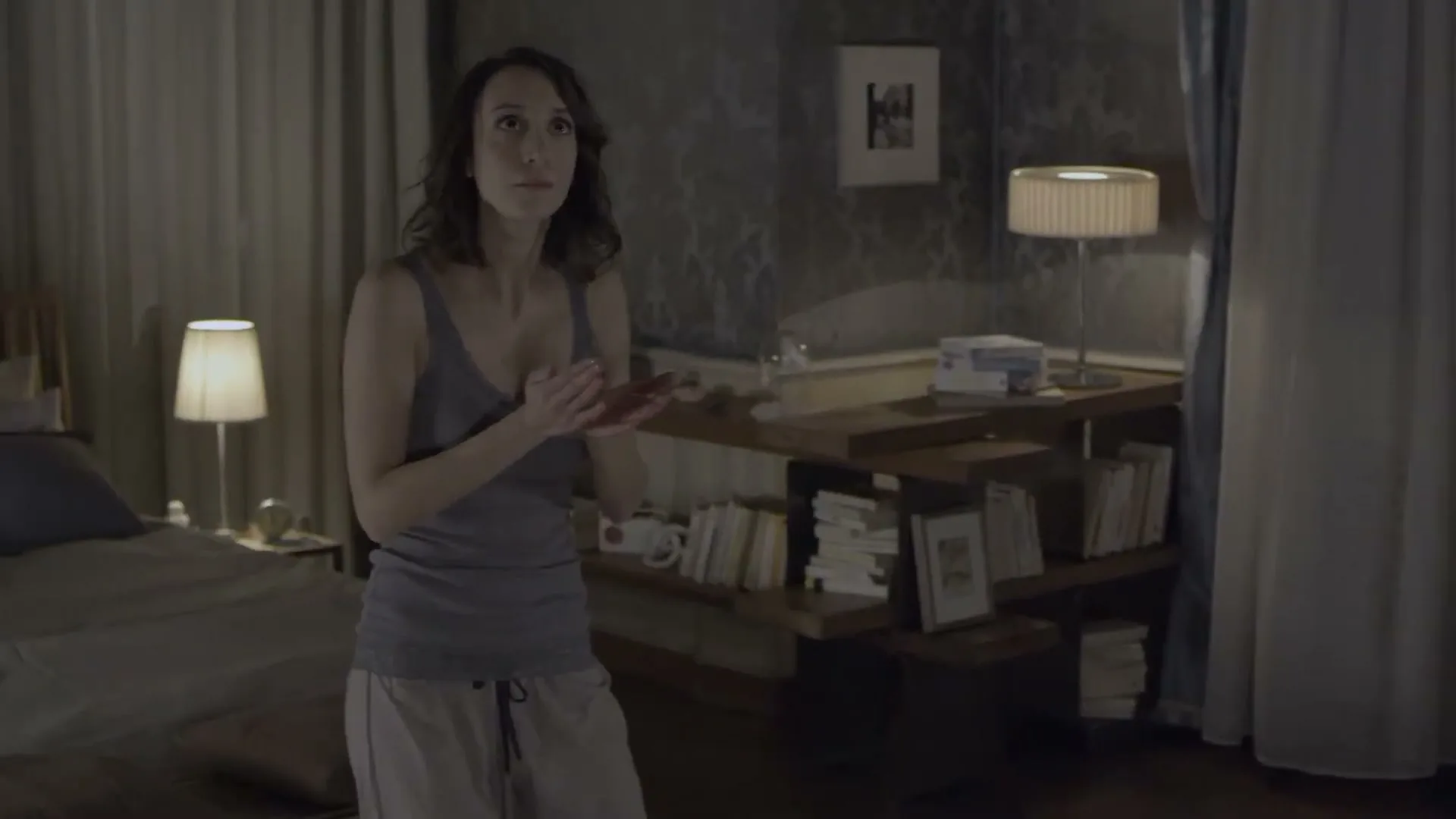 A woman in her 30s, wearing a grey tank top and light-colored pajama pants, stands in a dimly lit bedroom, looking around with a concerned expression.